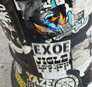A sticker featuring a blue duck character with sunglasses and wings, alongside stickers reading 'EXOE' and 'JIGLA FOO WITH A VENGEANCE', affixed to a black surface.