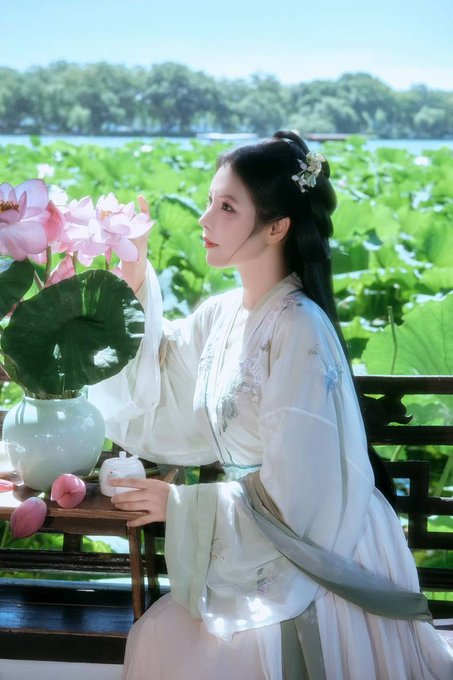 A young woman in traditional flowing hanfu-style clothing sits by a lotus pond, holding pink lotus flowers in a celadon vase.