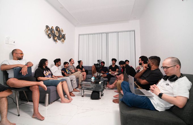 A group of people sits casually on gray sofas in a living room, gathered around a coffee table with laptops, phones, and cameras, appearing to watch a video.