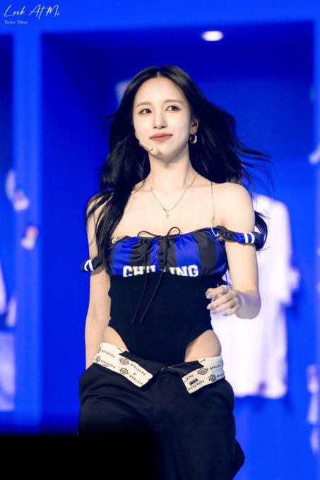 Mina of TWICE performs on stage in a black and royal blue corset-style top with "CHRISTIAN" partially visible across the front, paired with low-rise pants featuring branded waistband tape, looking stunning under blue stage lighting.