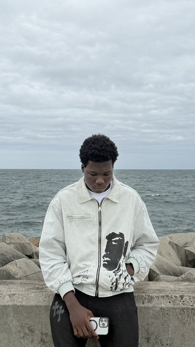 The image features a young man standing near a body of water, possibly an ocean or sea. He is wearing a white jacket with a black face design on the front. The man has his hands in his pockets while looking off into the distance. In the background, there are rocks and clouds, adding to the overall atmosphere of the scene. The post includes text that reads "good morning chase motion yap about $RIVER while you'r at it". This suggests a connection between the image and the caption, possibly indicating an ongoing chase or adventure related to the river depicted in the photo.