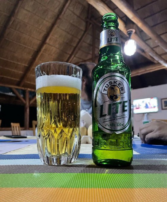 In the image, there are two glasses of beer sitting next to each other on a table. One glass has a green beer bottle behind it, while the other glass contains a white foam beer. The scene appears to be set outdoors or at an outdoor event, as suggested by the presence of a chair and dining table in the background. The image also includes a caption that reads "A very calm day like today demands such a sip.. #OwnTheLite." This message seems to be related to the beers being served, possibly promoting a brand or encouraging people to enjoy their time outdoors on a calm day with a refreshing drink.