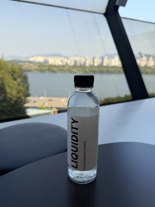 A bottle of 'LIQUIDITY' water by Ruckit sits on a dark table with a scenic view of water and a city skyline in the background.