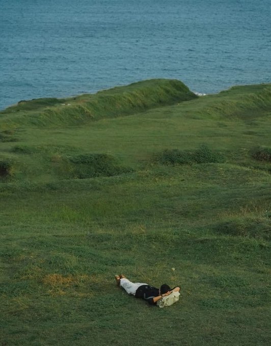 A person lies on a grassy coastal hillside overlooking the ocean, apparently napping with a bag beside them—ironically "touching grass" while the accompanying text jokes about missing their PC instead of enjoying nature.