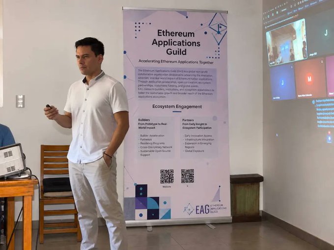 A man in a white shirt and light pants stands presenting next to a vertical banner titled "Ethereum Applications Guild" with the subtitle "Accelerating Ethereum Applications Together" and information about ecosystem engagement.