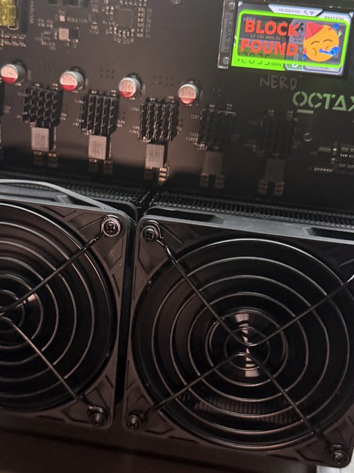 Close-up of a cryptocurrency mining rig featuring power supplies with cooling fans, circuit boards, and a small screen displaying a 'Block Round' sticker.