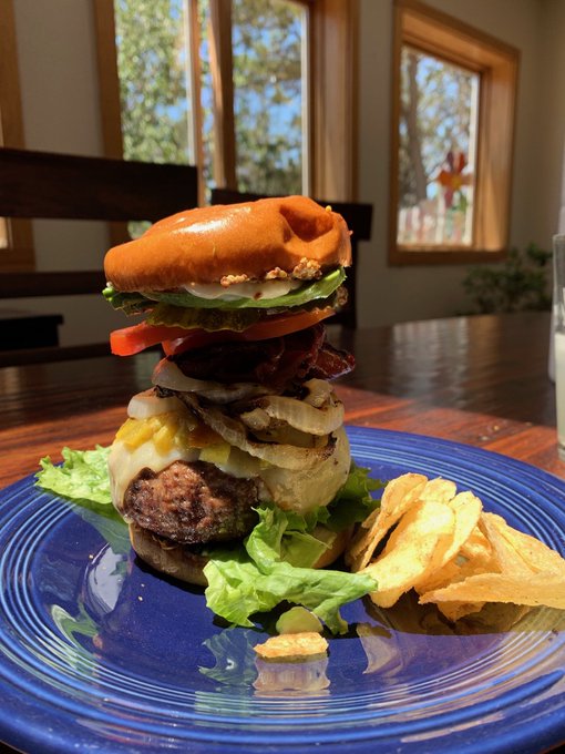 A towering homemade burger loaded with a beef patty, bacon, cheese, lettuce, and tomato sits on a blue plate alongside potato chips, photographed in a sunlit dining room.