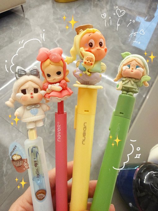 A hand holds four colorful pens topped with cute cartoon character figurines, including a bunny, a girl in a red dress, a baby, and a hooded figure, with some pens labeled 'never' and 'nusion'.