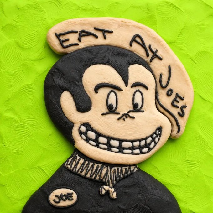 A clay sculpture of a cartoon character with a wide grin on a green background, with the text 'EAT AT JOES' and a name tag reading 'JOE'.