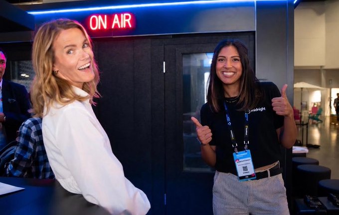 Two women smile inside a broadcasting studio under a neon 'ON AIR' sign; one wears a lanyard labeled 'midnight'.