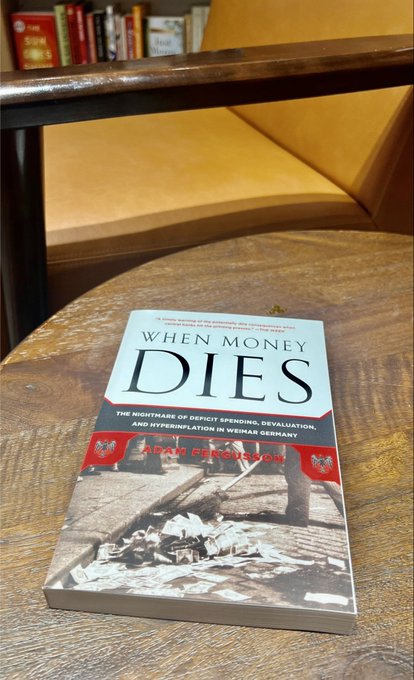 The book "When Money Dies: The Nightmare of Deficit Spending, Devaluation, and Hyperinflation in Weimar Germany" by Adam Fergusson rests on a wooden table next to a chair, with a quote praising it as "THE classic account of the economic and financial convulsions that struck Weimar Germany and their impact across Europe."