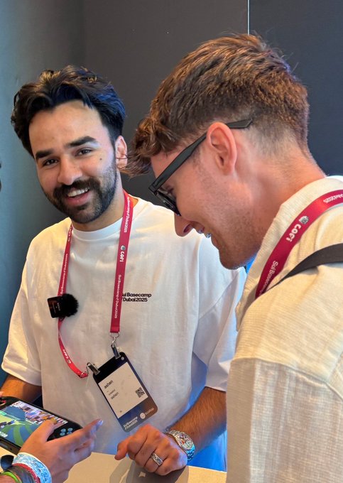 In this image, two men are standing next to each other at a desk with a computer monitor behind them. One of the men has a name tag that reads "Coffee Bean" and another man is wearing glasses. The man on the left side of the photo is holding a phone in his hand while looking at it. A post on social media shows an image of a Pokémon card, possibly related to the project they are discussing or the token they have received as a gift.