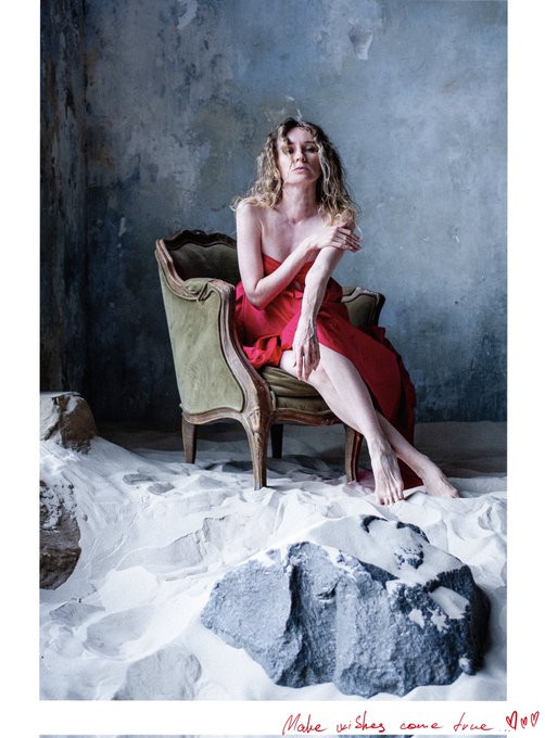 A woman with curly blonde hair in a red strapless dress sits barefoot on a green chair, posed against a textured blue wall and white fabric. Red text at the bottom reads 'Make wishes come true ♡♡'.