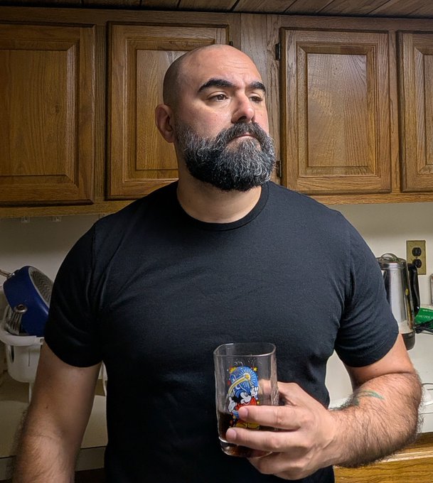Photo of a bald man with a thick grey beard wearing a black t-shirt, standing in a kitchen and holding a decorative glass with a colorful fish design.