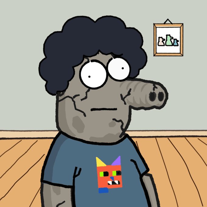 A cartoon character with dark curly hair, a cracked gray face, and a long snout wears a blue t-shirt featuring a colorful cat-like face design, standing in a room with wooden floors and a framed picture on the wall.