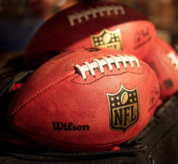 Several brown NFL footballs, branded with the "Wilson" name and the iconic NFL shield logo, are stacked together, visually representing the league's involvement in the legal battle over sports prediction markets.