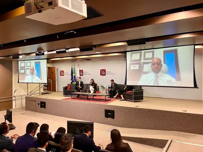 The image shows a panel discussion at the Brazilian Public Prosecutor's Council, where TRON's General Counsel John Hurston appears via video conference on large screens on either side of the stage, while seated panelists engage in conversation with an audience visible in the auditorium. The setup illustrates the post's description of a forum focused on digital asset recovery and consumer protection, highlighting the collaborative nature of the regulatory dialogue.