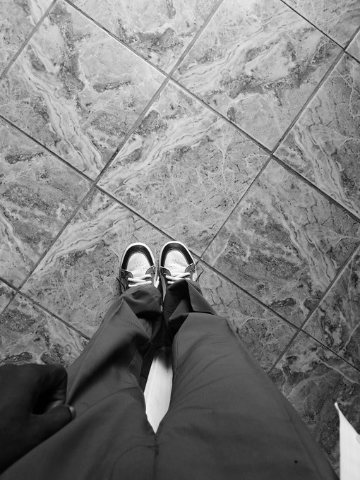 A black and white first-person perspective of someone looking down at their feet in sneakers standing on a marbled tile floor.