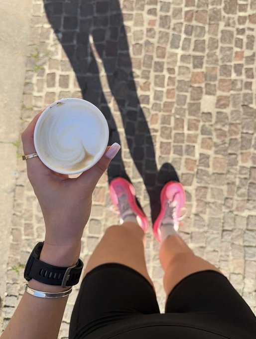 A first-person view looking down at someone's legs in black shorts and pink sneakers, holding a nearly empty coffee cup over a cobblestone street in bright sunlight.