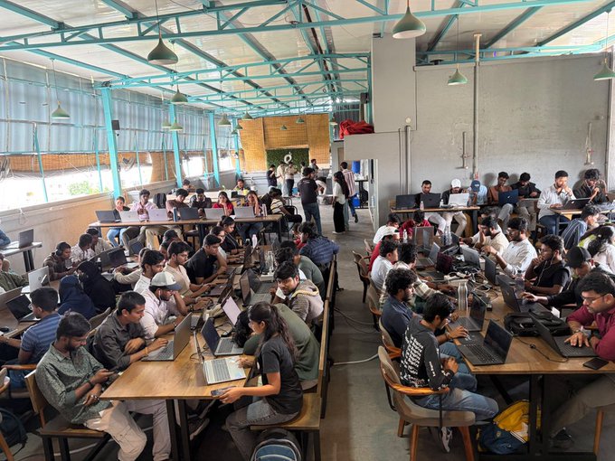 A large, crowded open-plan co-working space or classroom with blue ceiling beams, filled with people working on laptops at long wooden tables.