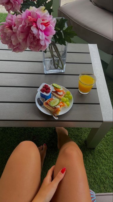 First-person view of someone relaxing outdoors with a breakfast plate (toast, vegetables, yogurt, fruits) and orange juice on a wooden table beside pink peonies.
