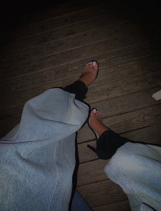 A first-person nighttime photo showing a person's feet wearing clear-strap heels with pink toenails, paired with light blue jeans on a wooden deck.