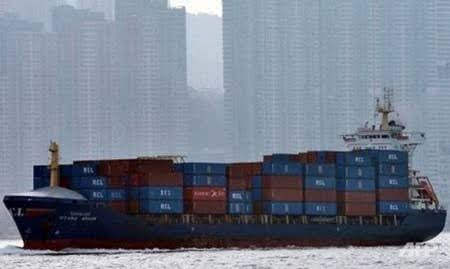 A large container ship loaded with multi-colored shipping containers sails through a harbor, with a hazy city skyline of high-rise buildings visible in the background.
