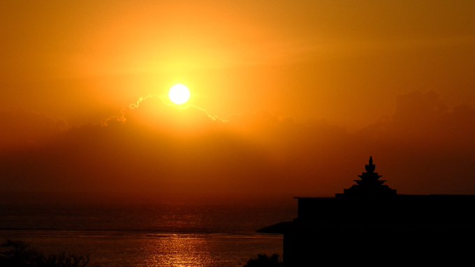 A vibrant sunset over the ocean features the dark silhouette of a building or structure in the foreground against an orange and yellow sky.