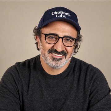 A smiling man with glasses and a graying beard wears a navy baseball cap embroidered with "Chobani Labs." The image aligns with the caption's entrepreneurial story of humble beginnings leading to a bold business venture, visually representing the founder's journey from grocery store apprentice to aviation investor.