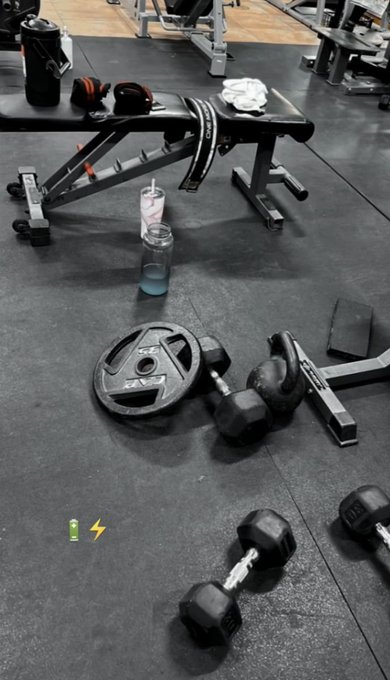 A gym scene showing a weight bench with dumbbells, water bottles, and headphones on a rubber floor, with weight plates scattered around.
