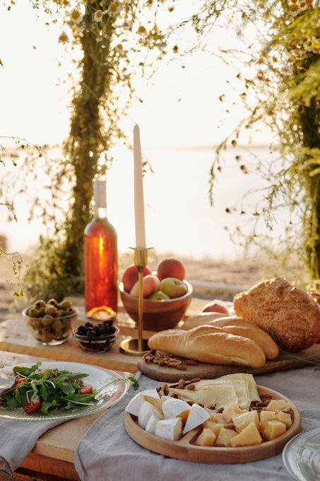 An outdoor table setting featuring an assortment of cheeses, bread, fruits, olives, greens, a bottle of rosé wine, and a lit taper candle at sunset.