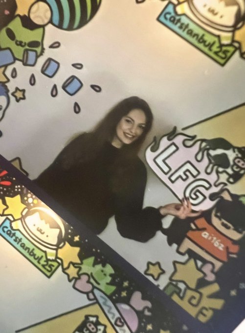 A slightly blurry, angled photo of a woman in dark clothing smiling and holding up a LEGO sign, standing in front of a colorful backdrop featuring various cartoon characters and the text 'catstanbul 25'.