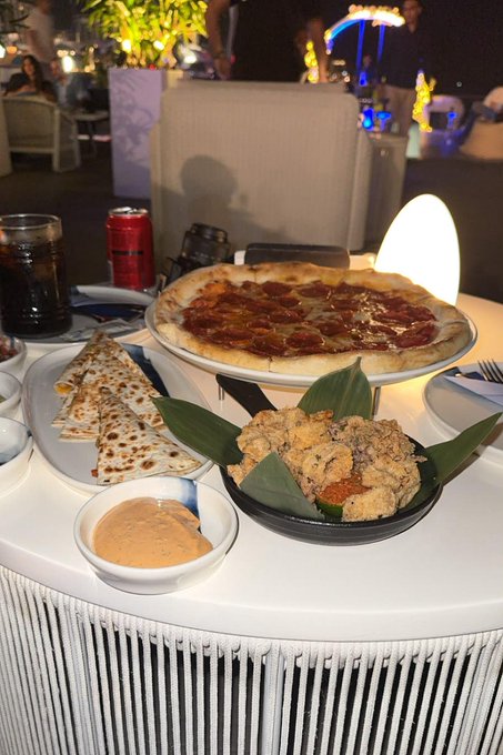 A table displays a pepperoni pizza, fried chicken bites in a leaf-lined bowl, and a small dish of sauce, all on a white round table at an outdoor restaurant. The variety of food shown suggests the "$4" price mentioned in the caption likely refers to the $SOL cryptocurrency payment for the meal rather than the cost, questioning whether the user was overcharged in crypto for this modest spread.