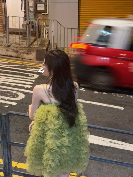 A woman with long dark hair wears a voluminous lime-green tulle dress while standing on a Hong Kong street as a red taxi speeds past, her back turned to the camera. The image visually captures the "Flashin' Hong Kong" moment from the post, showcasing her stylish arrival in the city courtesy of the AIW3 airdrop reimbursement.