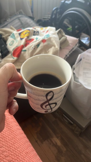 A hand holds a white ceramic mug decorated with a treble clef and musical notes, filled with black coffee.