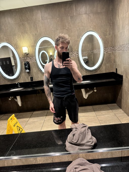 A bearded man takes a mirror selfie in a public restroom while wearing a black tank top and athletic shorts, perfectly capturing the "GM" (Gym Morning) vibe of an early workout session. A yellow "CAUTION" wet floor sign sits on the tiles behind him, and a towel rests on the counter in the foreground.