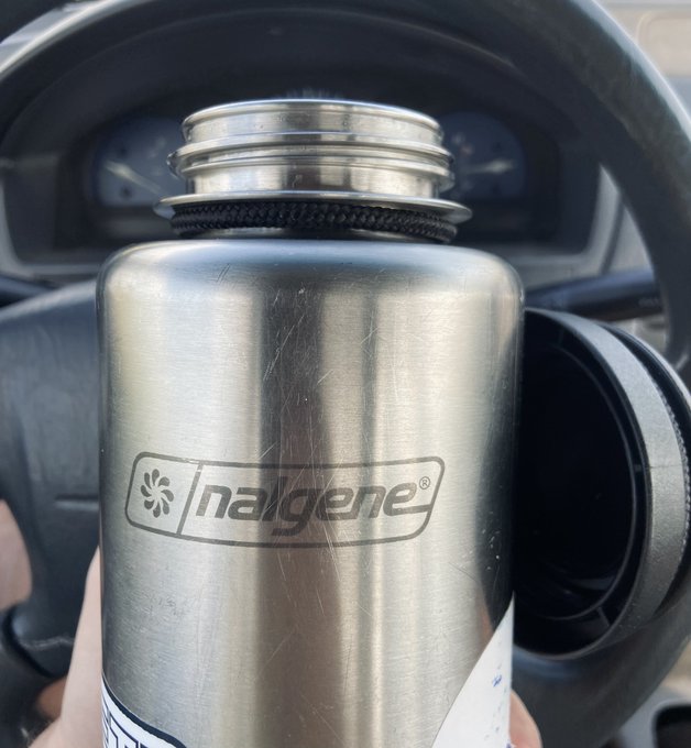 A hand holds a stainless steel Nalgene bottle in front of a vehicle's steering wheel and dashboard, highlighting its vintage-style metal design with the prominent "nalgene" logo—a rare find referenced as "a true grail" among collectors.