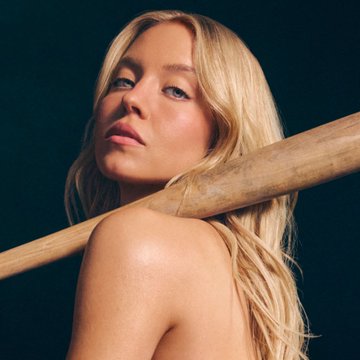 A blonde woman with long, wavy hair poses bare-shouldered against a dark background, with a wooden pole or bat resting across her neck and shoulder in a fashion-style shot. The image appears to be promotional content, likely referencing Sydney Sweeney's recent advertising controversy involving athletes critiquing her nearly nude campaign for a sports prediction platform.