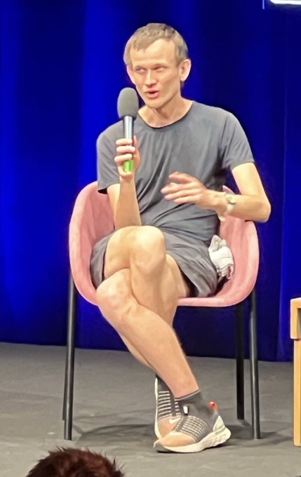 A man in a gray t-shirt and shorts sits cross-legged on a pink chair, holding a microphone while speaking on a stage with a blue curtain backdrop. The image captures a casual yet engaged speaker, with the caption "unreal levels of bullish" suggesting his words convey strong optimism or enthusiasm.