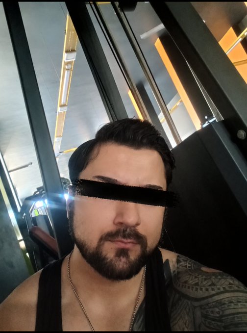 A selfie of a bearded man with a black censor bar over his eyes, wearing a black tank top and showing a tattooed shoulder, taken in what appears to be public transportation.