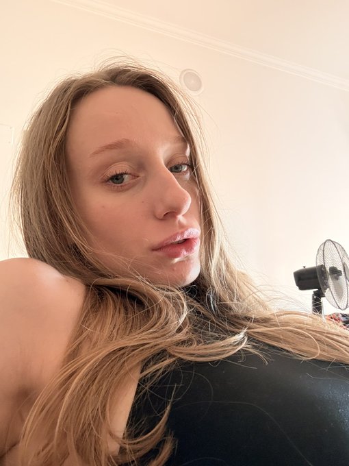 A close-up selfie of a young woman with long blonde hair looking down at the camera, with a small fan visible in the background.