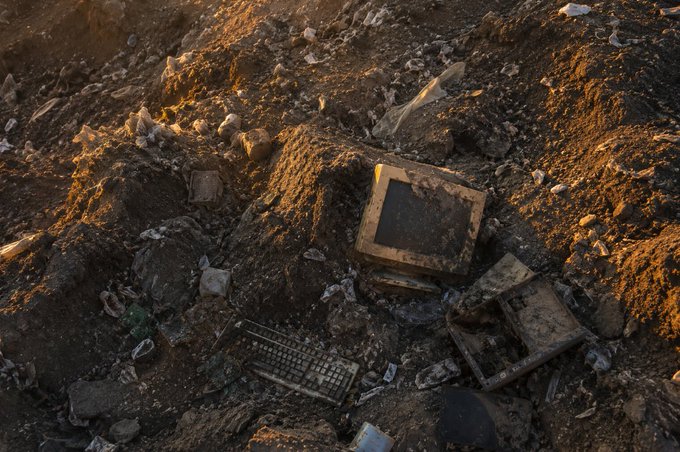 Discarded computer monitors and a keyboard half-buried in a pile of dirt and rubble, illuminated by golden sunset light.
