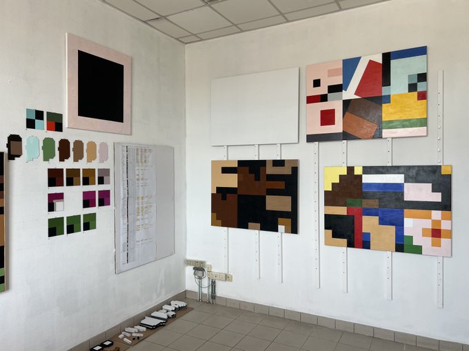 A studio gallery displays abstract geometric paintings featuring bold color blocks and grid compositions in earthy and primary tones, with color swatches mounted on the left wall and sketches/papers arranged on the floor. The "Gm" greeting pairs playfully with the art space's creative, morning-fresh atmosphere of artistic exploration and palette studies.