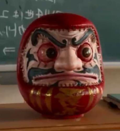 A traditional red Daruma doll with a fierce painted face, featuring thick black eyebrows, wide eyes, and gold decorative patterns on its base, sitting in front of a chalkboard.