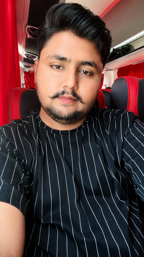 A selfie of a man with dark hair, a mustache, and a short beard, wearing a black shirt with thin white vertical stripes, seated inside a vehicle with red seats and red curtains.