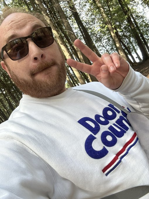 A selfie of a bald man with a beard wearing sunglasses and a white sweatshirt with 'Doof Cous' text, making a peace sign in a forested area.