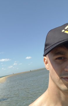 A shirtless man wearing a black cap with yellow lettering poses for a selfie at a serene beach, where shallow turquoise waters meet a clear blue sky. The image pairs perfectly with the "#Binance" theme, suggesting a relaxed getaway that blends crypto enthusiasm with a beautiful coastal escape.