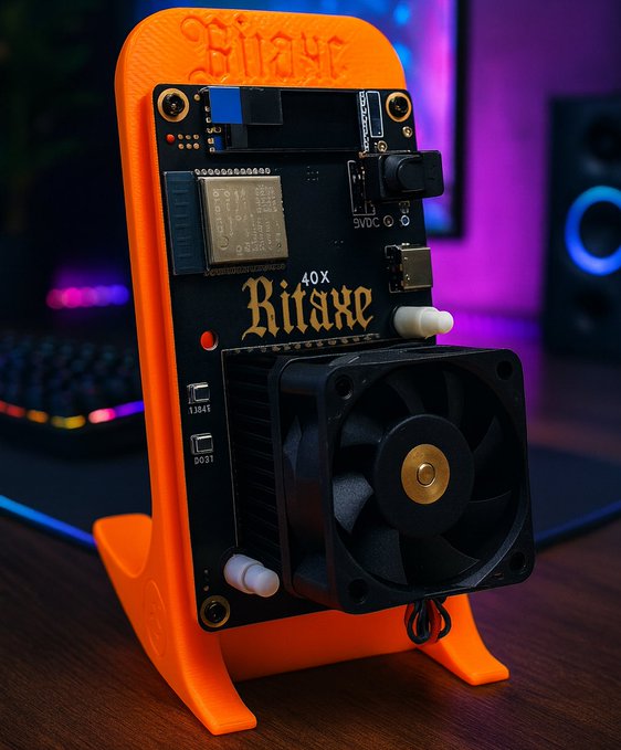 A Bitaxe solo miner (labeled "40X Bitaxe") stands upright on an orange 3D-printed stand, featuring an exposed circuit board, a single ASIC chip, and a cooling fan on a gaming desk setup. The image illustrates the single-chip Bitaxe device referenced in the post, which mined block 957,382 to claim a 3.1382 BTC reward.