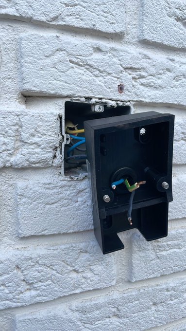 A black electrical fixture is mounted on a white brick wall, with wires visible in the wall cavity behind it.