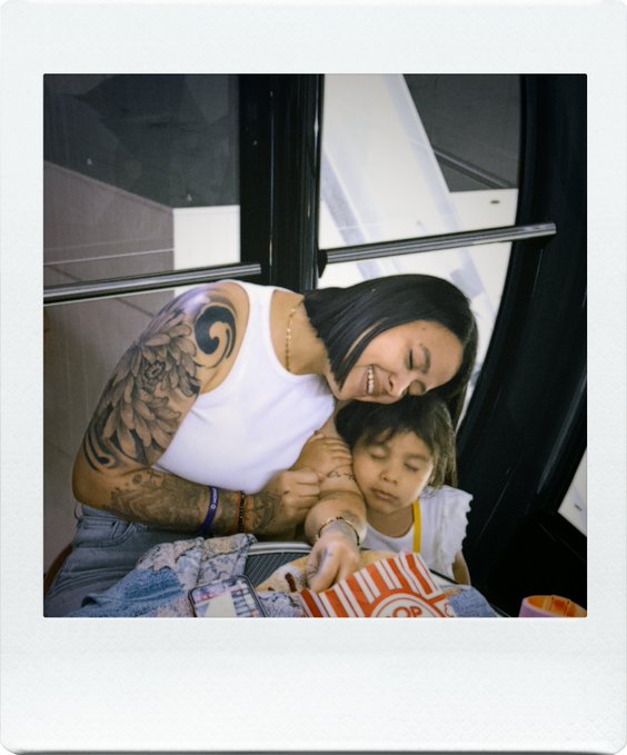 A polaroid-style photo of a woman with short black hair and a large arm tattoo, wearing a white tank top, smiling warmly as she cuddles with a young child who appears to be asleep in her lap, holding what looks like a container of popcorn.